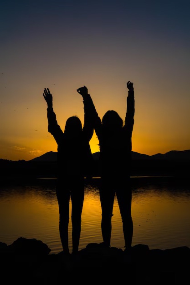 Two friends silhouetted at sunset, arms raised