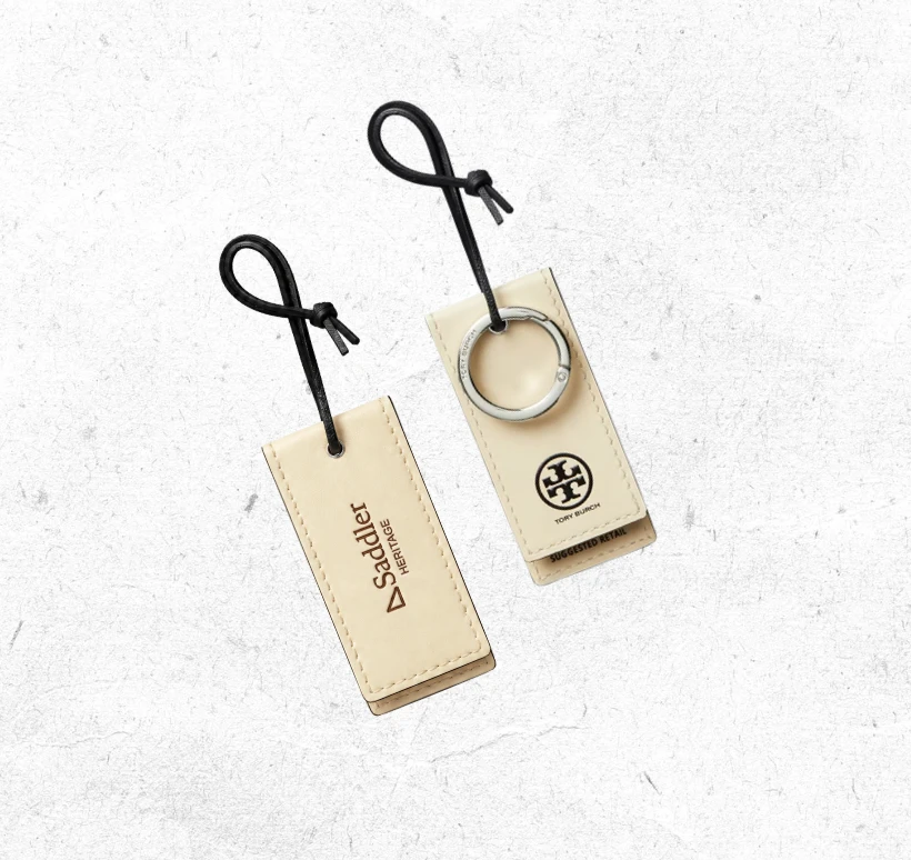 Saddler hang tags in cream leather: debossed Saddler Heritage wordmark on one, Tory Burch logo on the other